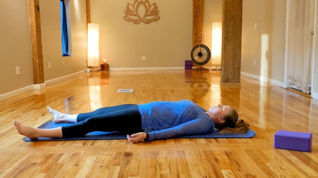 Sam lies on a purple yoga mat in a yoga studio in savasana pose.