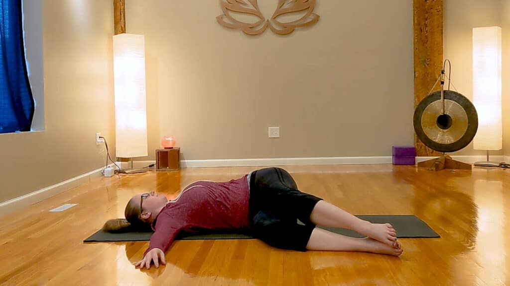 Sam is lying on a black yoga mat in a reclined twist pose. She is wearing black capri leggings and a pink jacket.
