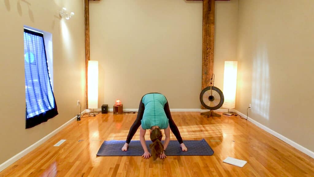 Sam is standing in a wide legged forward fold on a purple yoga mat. She is wearing black legggings and a teal shirt.