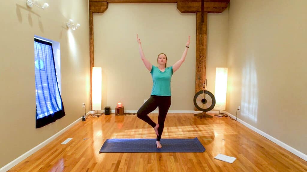 Sam is standing on a purple yoga mat in tree pose with her arms overhead. She is wearing a teal shirt and black leggings.