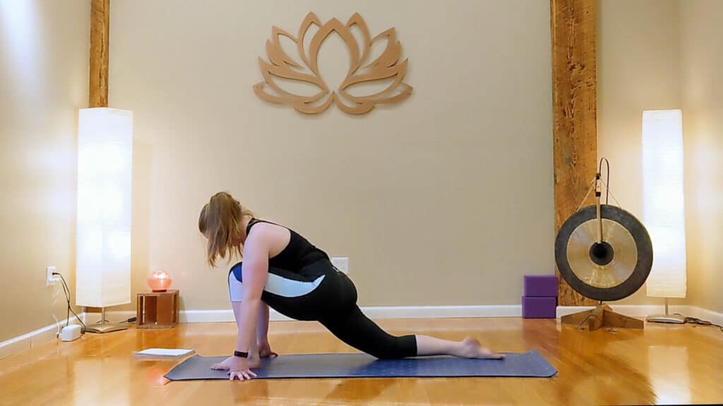 Sam is in low lunge pose on a purple yoga mat. She is wearing a black tank top and black carpis with a white stripe on the side.